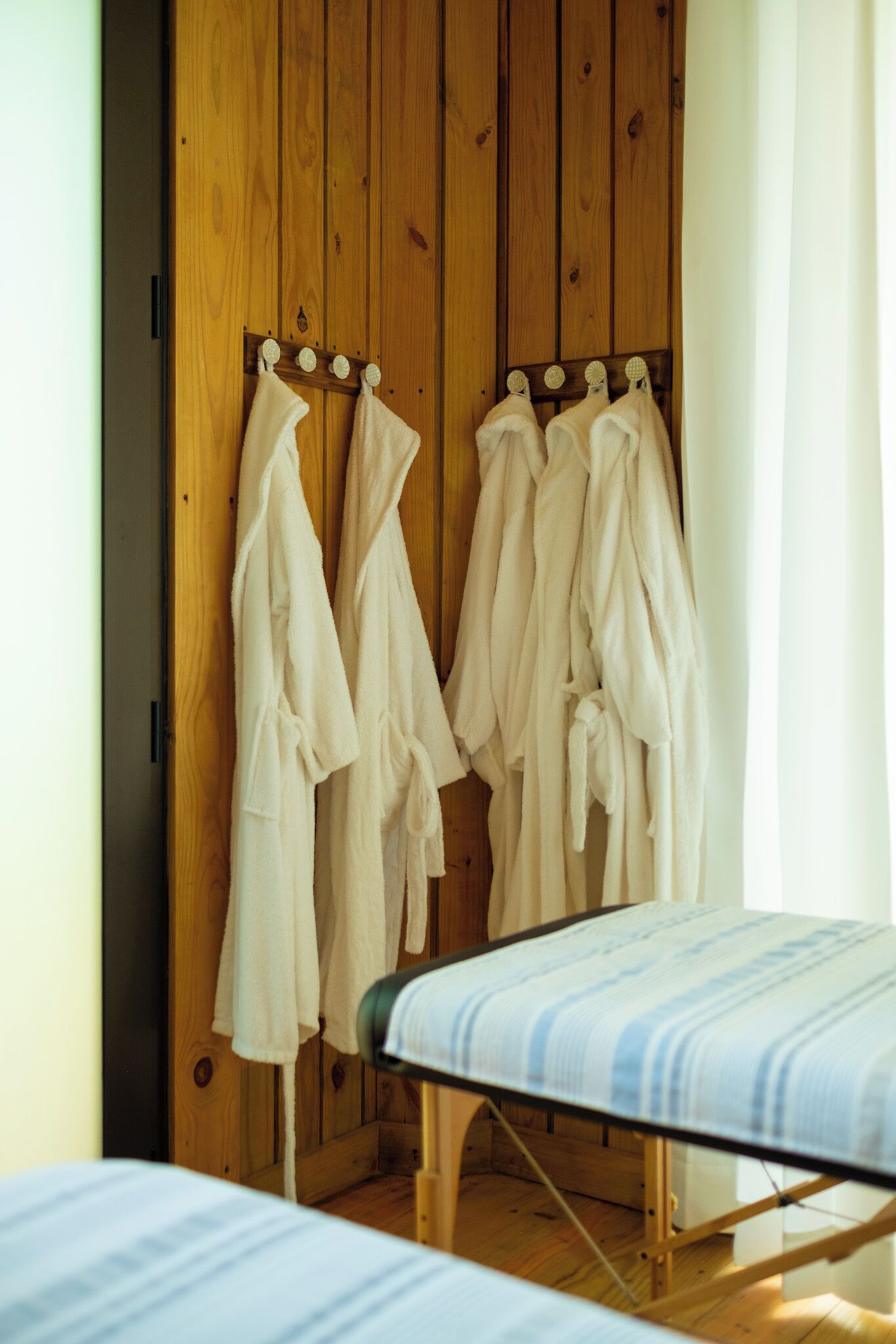 A wooden wardrobe with multiple hanging robes and a bed with a striped bedspread.