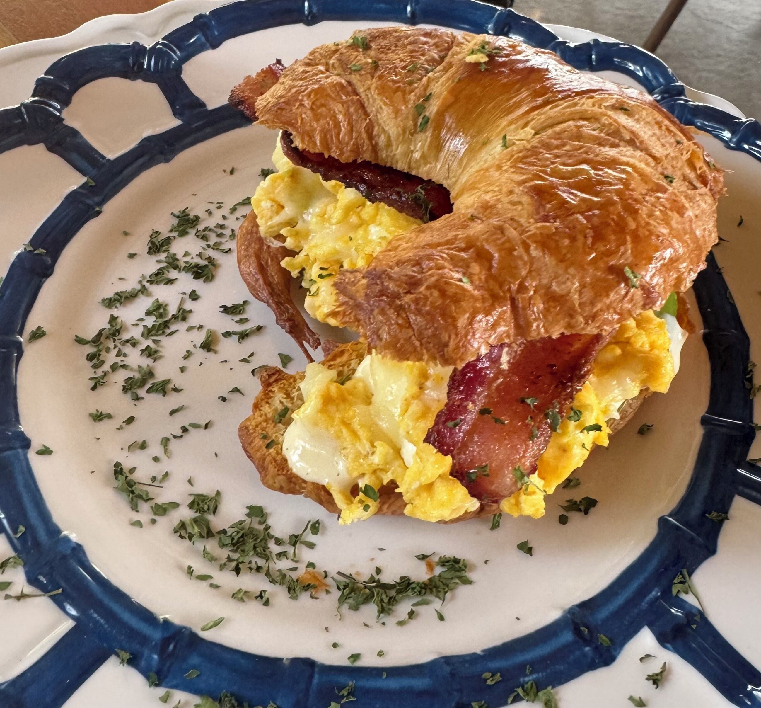 Ein Croissant-Sandwich mit Rührei und Speck auf einem Teller mit blau-weißem Muster.