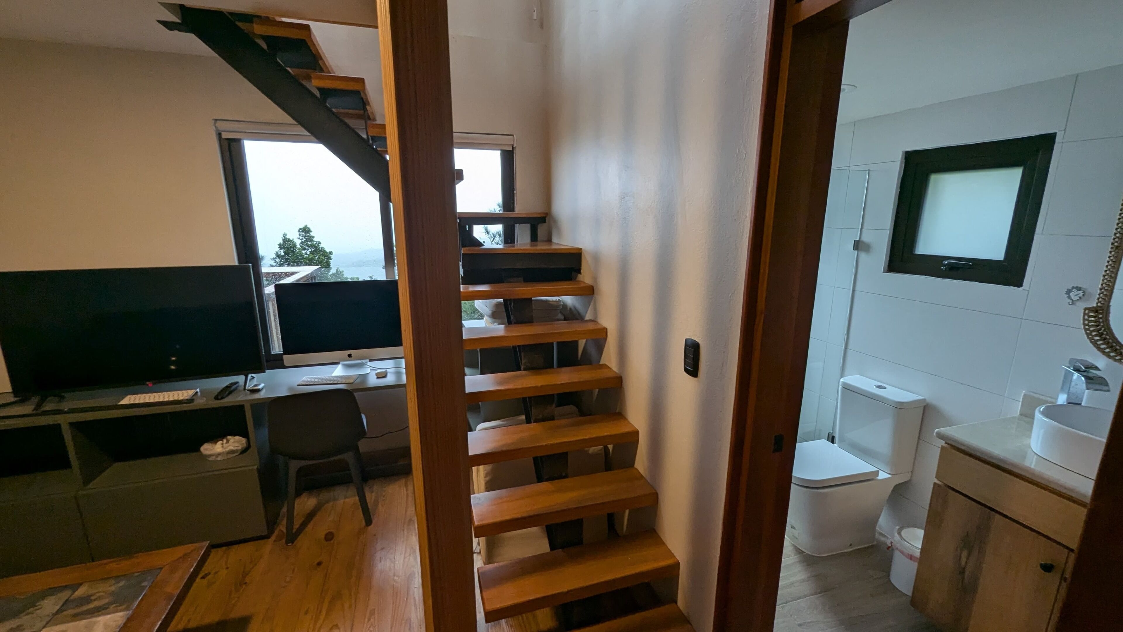A room with a wooden staircase, a desk with a computer, a television, and a bathroom with a toilet and sink.