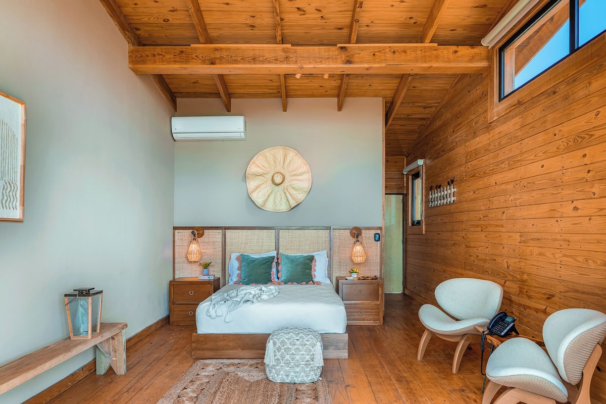 A bedroom with a wooden ceiling, a large round wall decoration, a bed with pillows, bedside lamps, a bench, and two chairs.