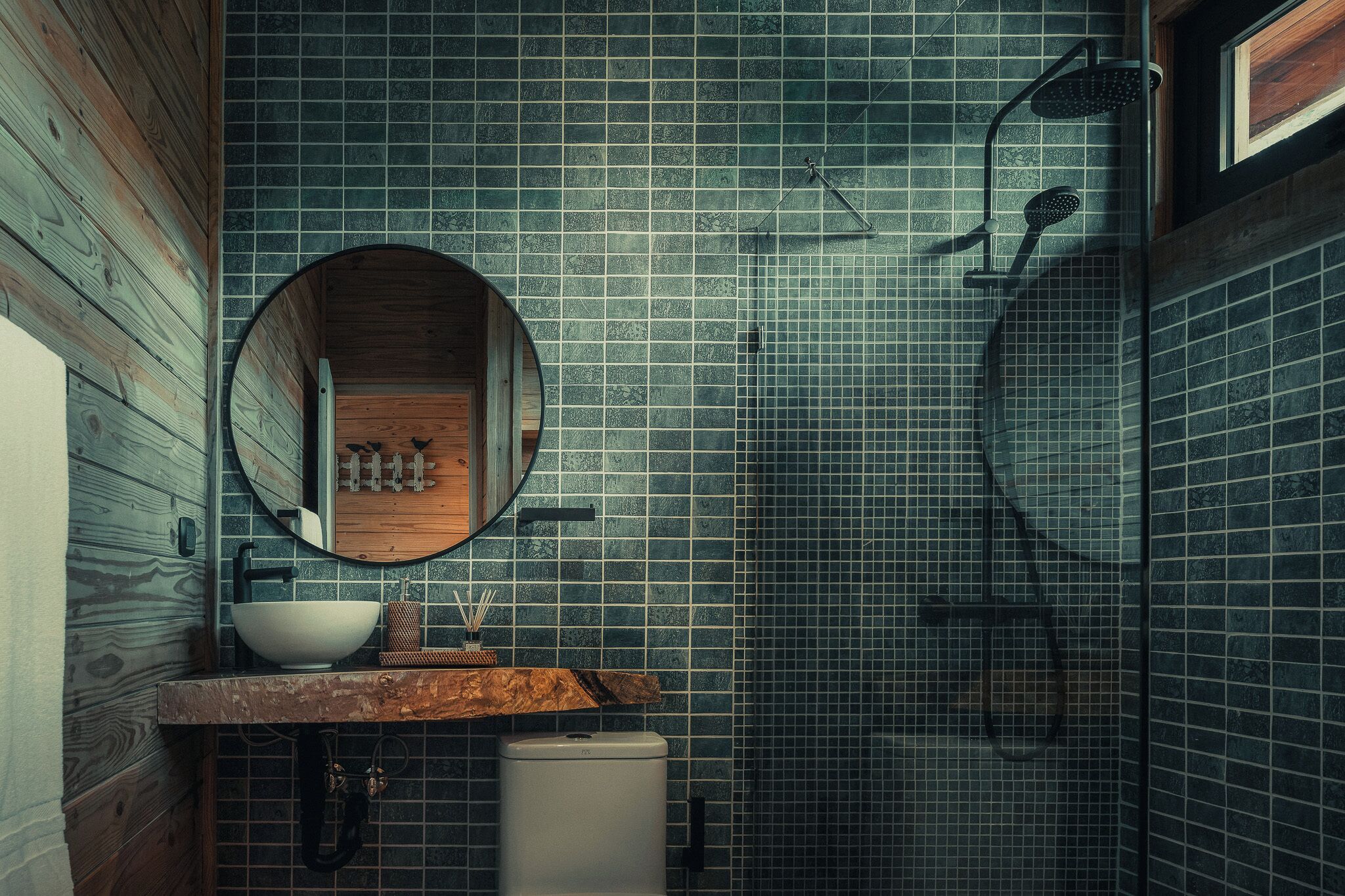 Un baño con tocador de madera, un espejo redondo, un lavamanos blanco y una zona de ducha.