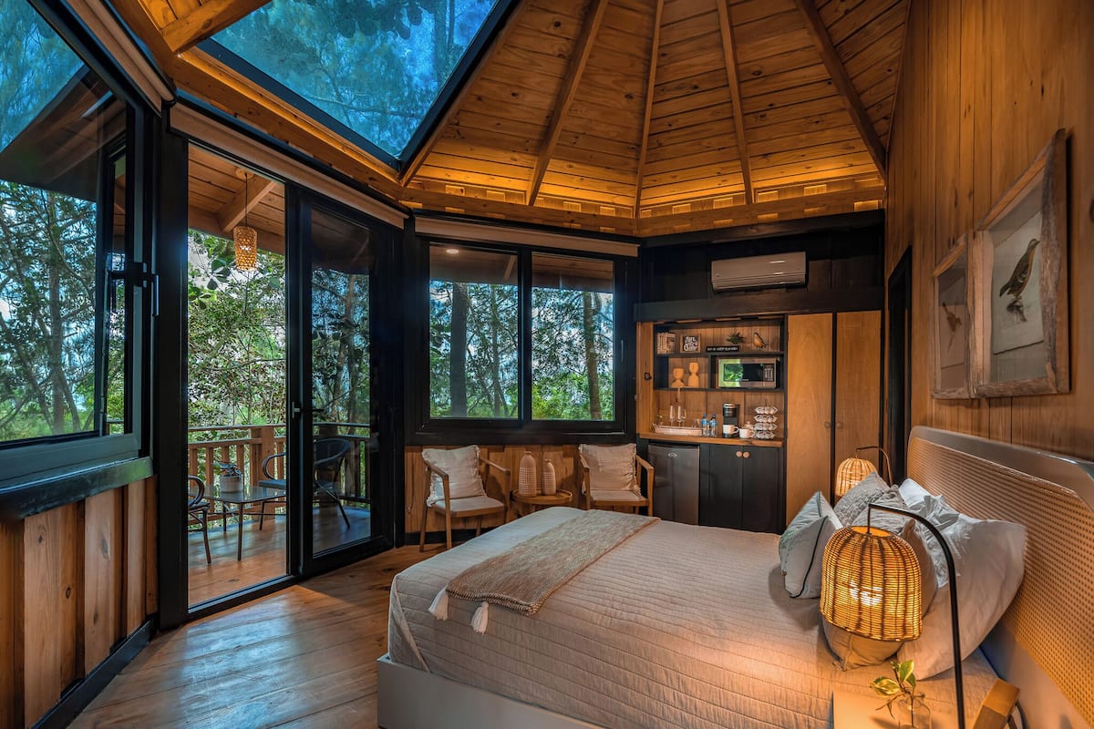 A spacious bedroom with a large bed, a wooden ceiling, and a view of the forest through large windows.