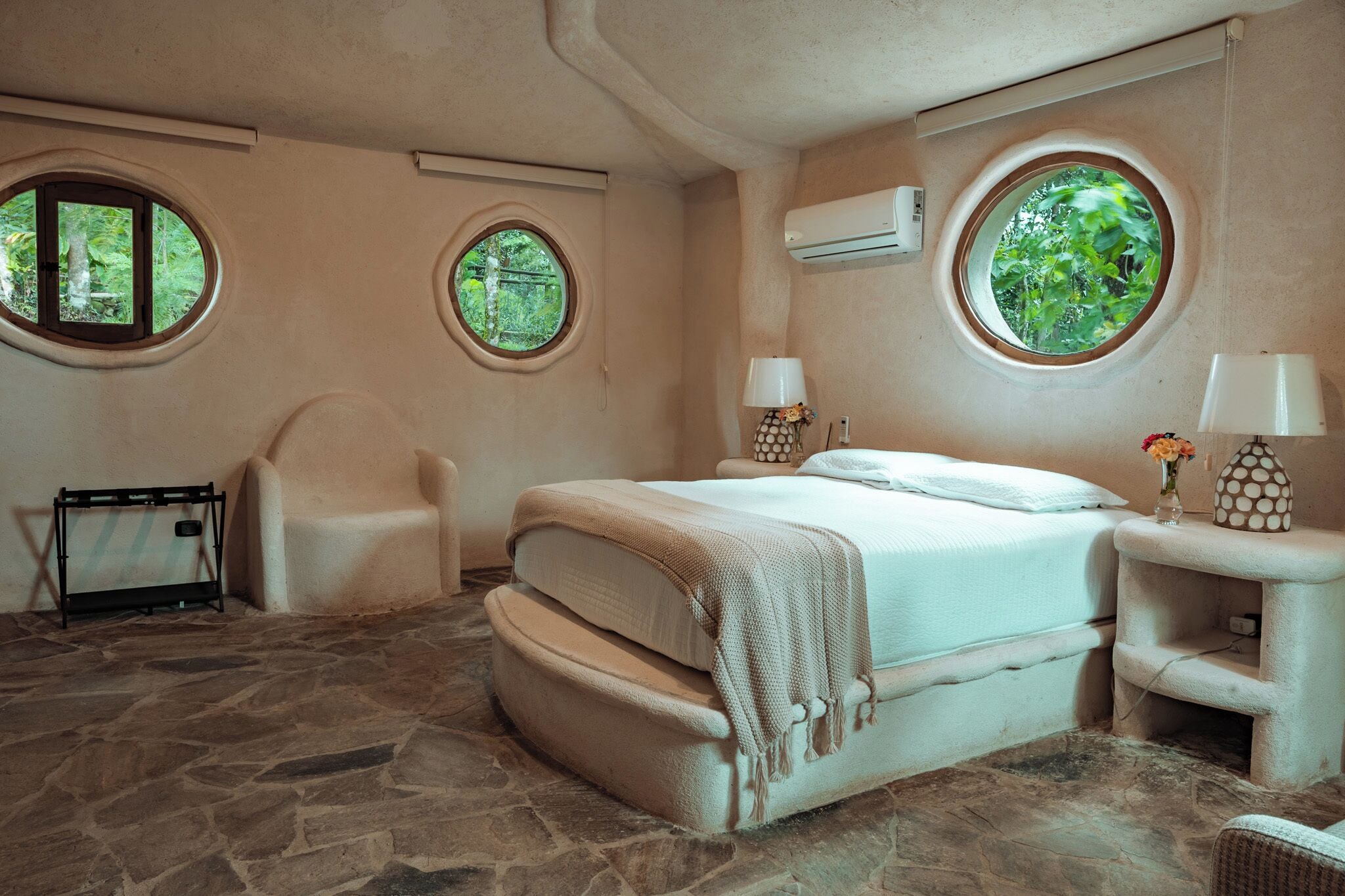A bedroom with a stone floor, a bed with a beige blanket, two round windows, a chair, a nightstand with a lamp, and an air conditioning unit.
