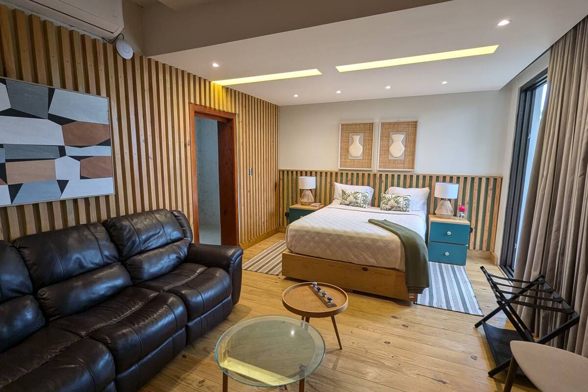 A modern bedroom with a leather sofa, a glass coffee table, a bed with a wooden headboard, and a nightstand with lamps.