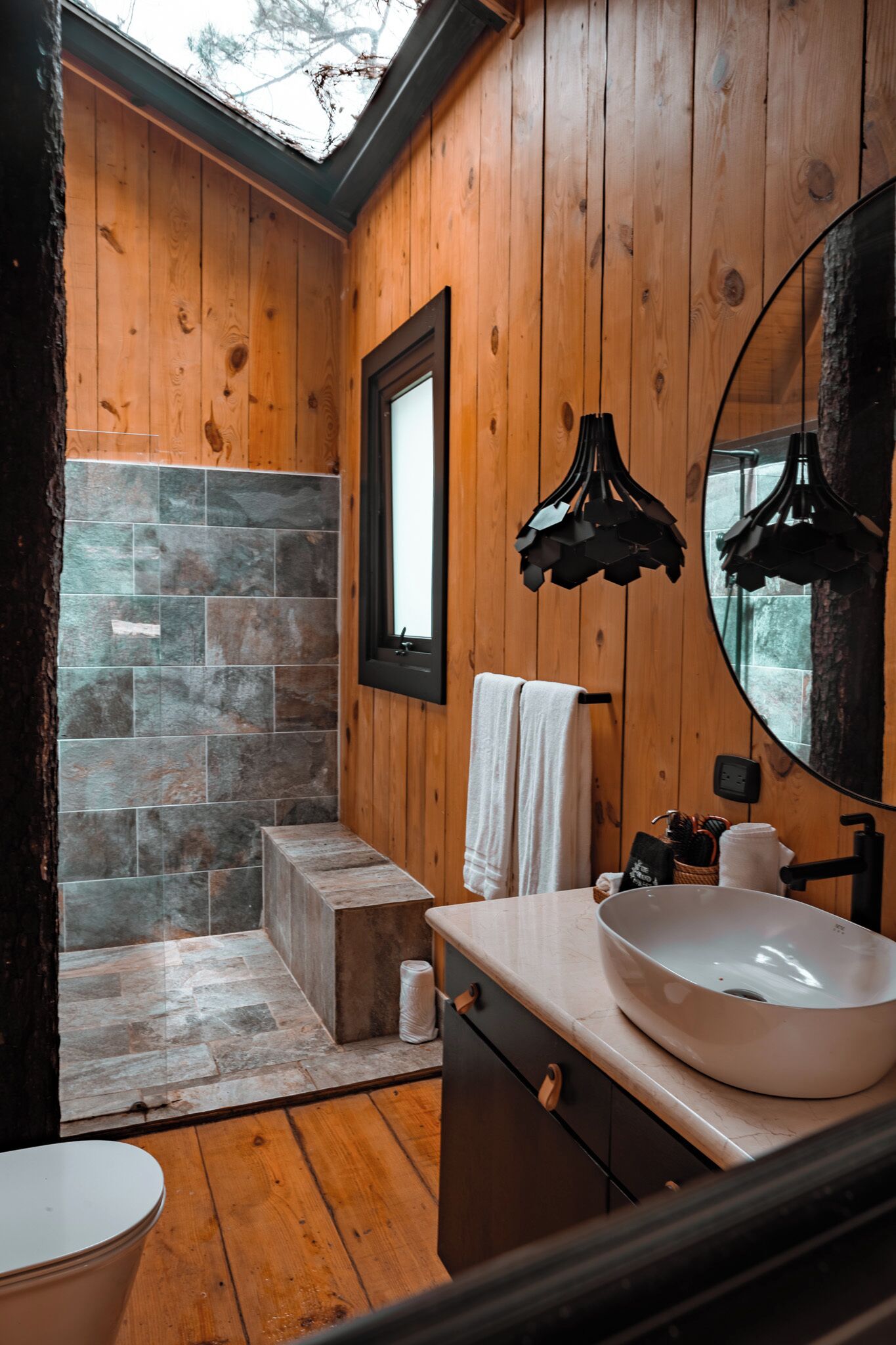 A wooden-paneled bathroom with a stone shower, a large mirror, and a white sink.
