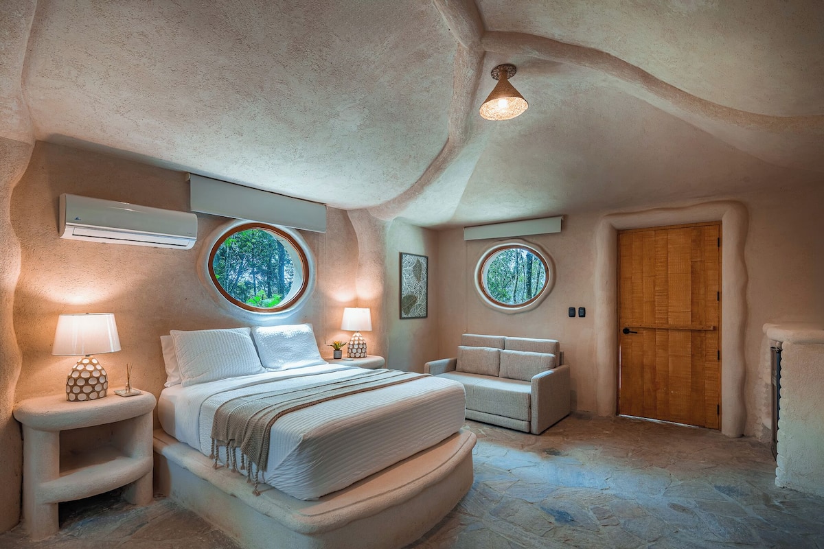 A bedroom with a large bed, two bedside tables with lamps, a wooden door, and two round windows with views of greenery.