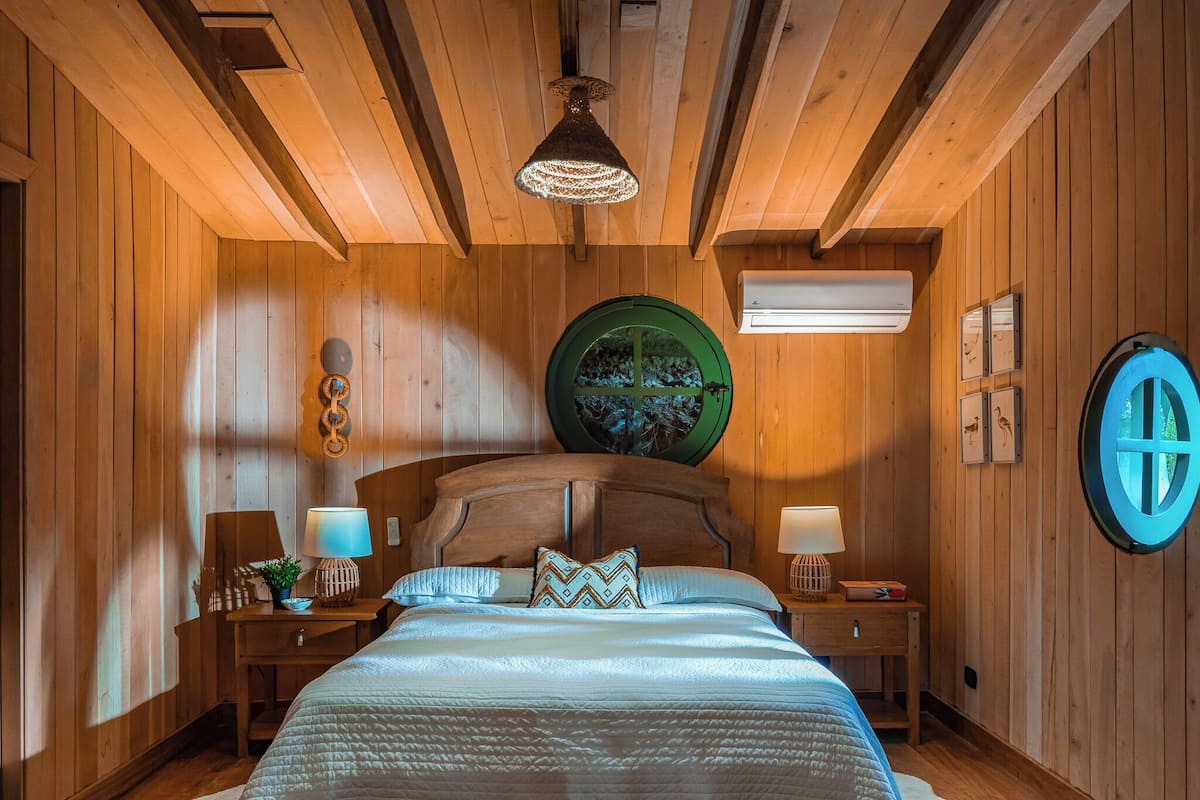 A wooden-paneled bedroom with a large bed, two bedside tables, a wall-mounted circular window, and a ceiling light fixture.