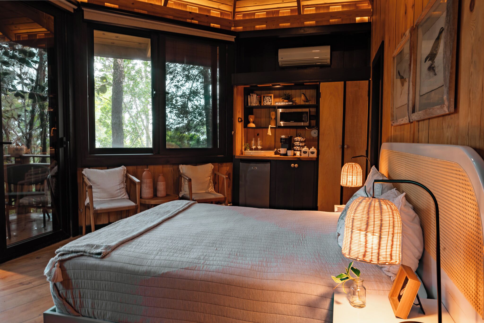 A cozy bedroom with a large bed, a bedside table with a lamp, a small kitchenette, and a seating area with chairs.