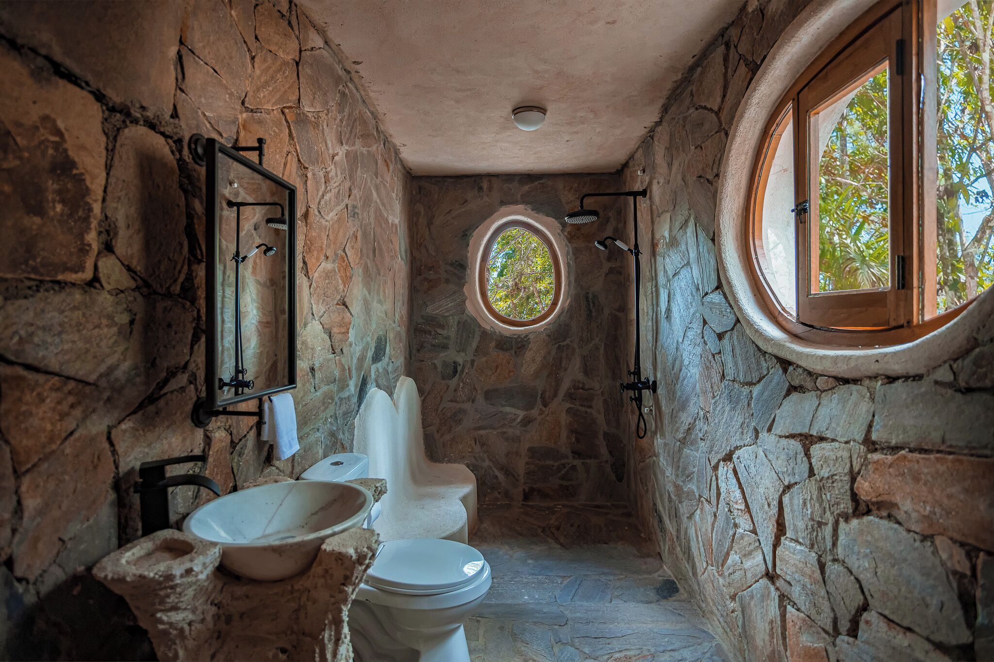 A stone-walled bathroom with a round window, a white toilet, and a basin.