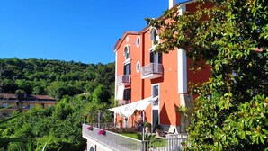 A multi-story building with balconies, a terrace with outdoor seating, and lush greenery in the background.