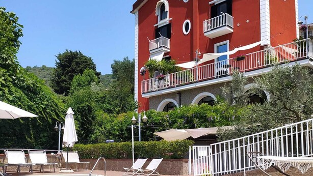 A swimming pool with lounge chairs and umbrellas, surrounded by a red building with balconies and a garden.