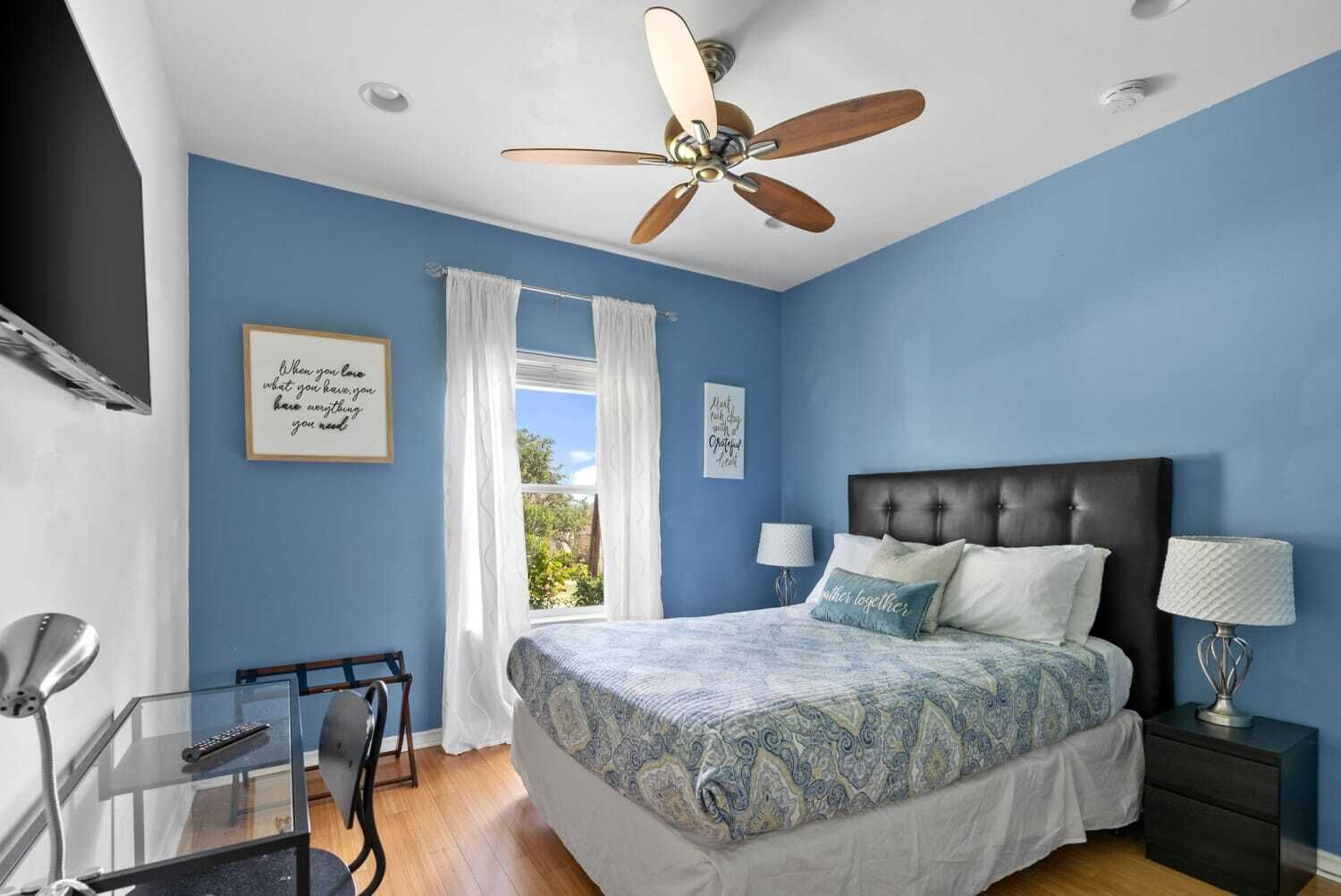 A bedroom with a bed, a ceiling fan, a window with curtains, a bedside table with a lamp, and a wall with a framed quote.