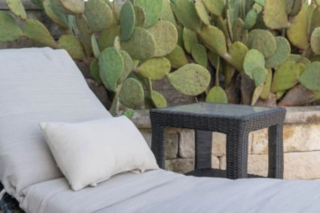 Een bed met witte beddengoed en een kussen, een klein zwart rieten tafeltje, en een cactus in de achtergrond.