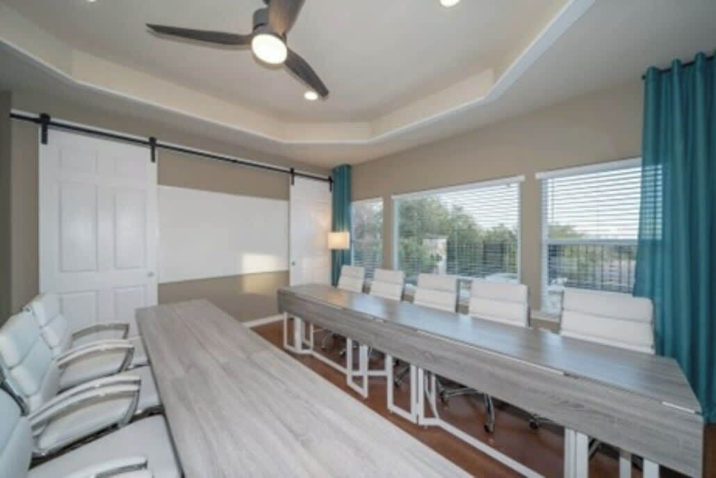 A conference room with a long table, white chairs, a ceiling fan, and large windows with curtains.