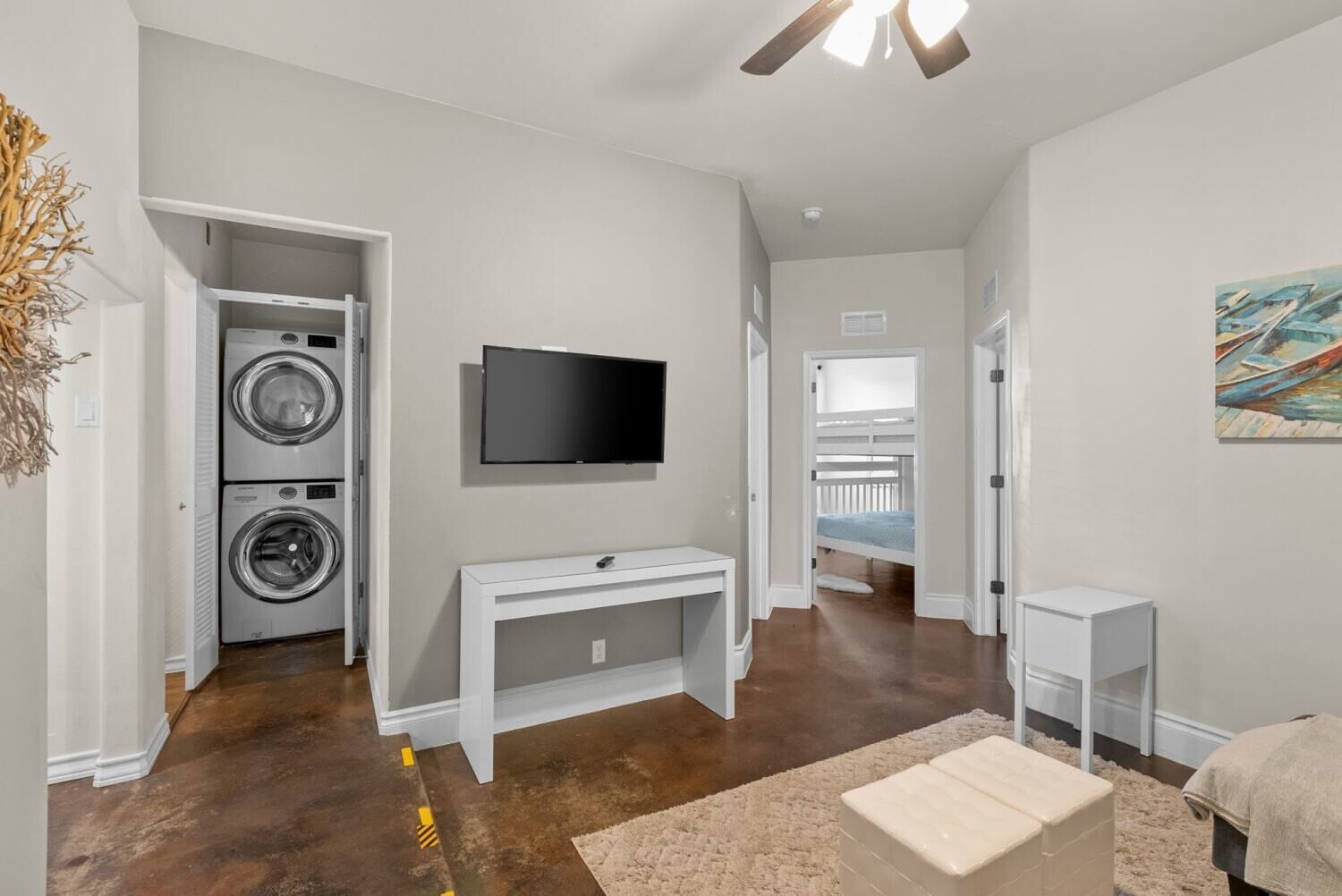 Une buanderie avec des lave-linge et des sèche-linge superposés, un téléviseur à écran plat fixé au mur, une console blanche et, au fond, un lit superposé.