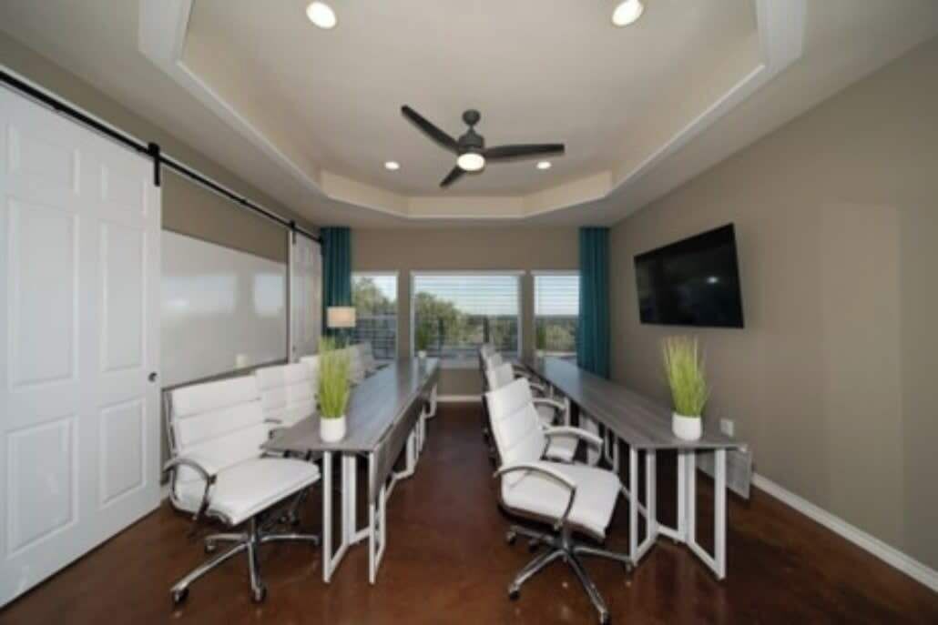 A conference room with a long table, white chairs, a ceiling fan, and a TV on the wall.