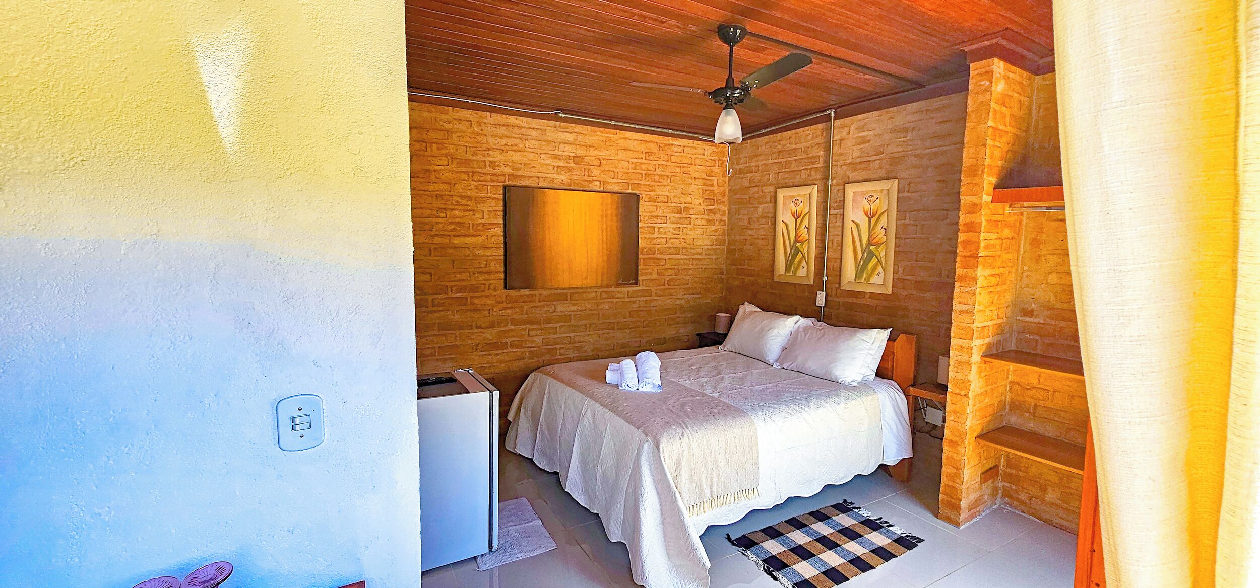 Une chambre à coucher avec un lit, un ventilateur de plafond, un téléviseur fixé au mur et un mur en briques.