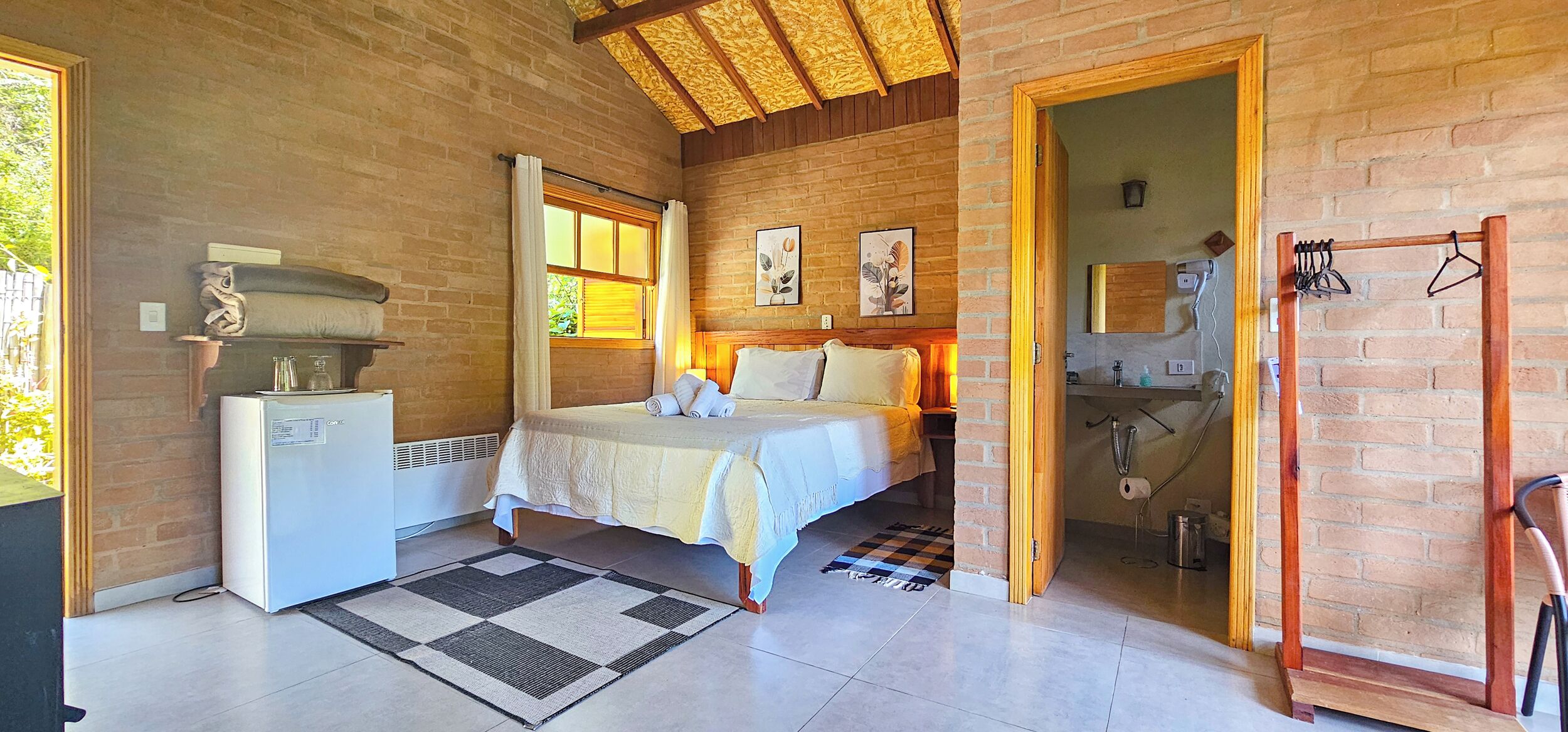 Une chambre avec un lit, un petit réfrigérateur, une porte en bois donnant sur une salle de bain et un tapis à carreaux.