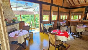 A rustic dining area with multiple tables, each with a red tablecloth and white table runners. The room features exposed wooden beams and brick walls, with large windows providing views of greenery outside.