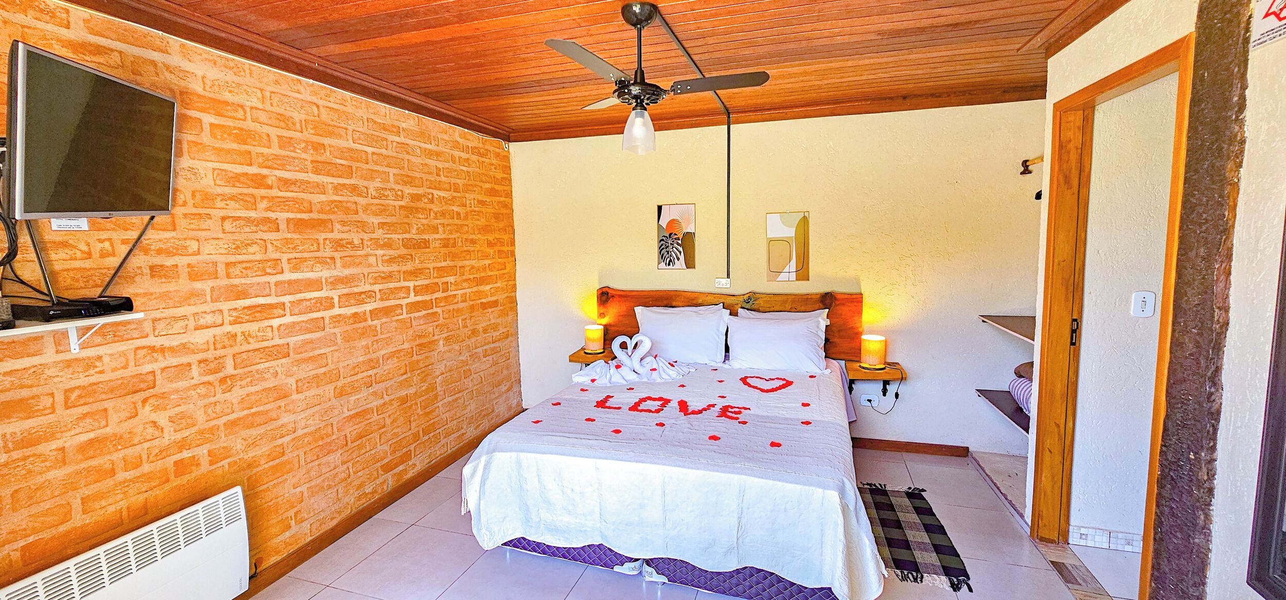 A bedroom with a brick wall, a bed with a 'LOVE' decoration, a TV, and a ceiling fan.