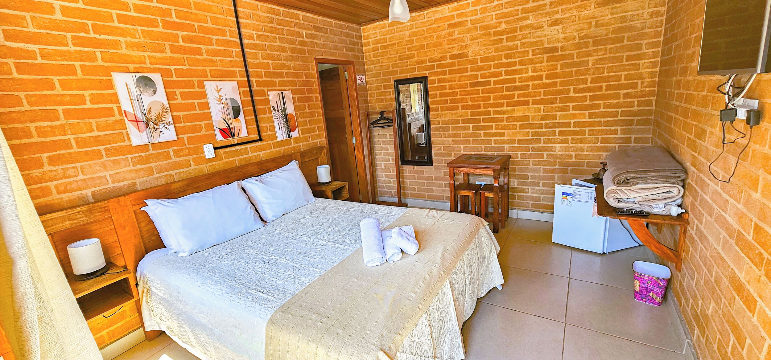 A room with a bed, a wooden nightstand, a small table, a chair, a mirror, and a wall-mounted TV.