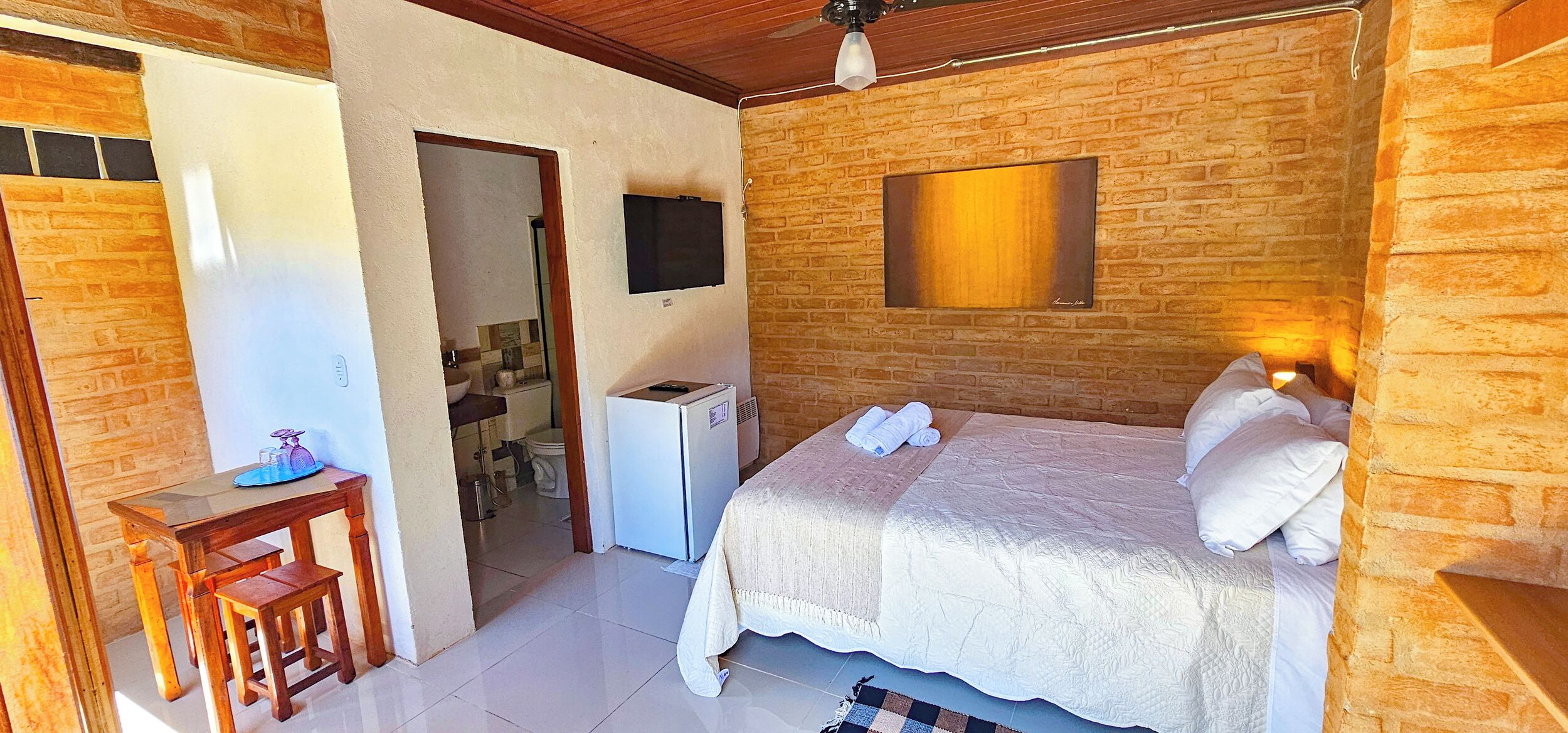 Un dormitorio con pared de ladrillo, una cama con ropa blanca, una mesita con lámpara y un televisor montado en la pared.