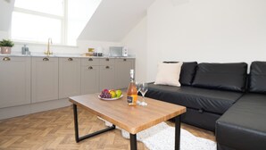 A modern kitchen with grey cabinets, a wooden table with a bottle and glasses, and a black sofa in a bright room with a window. - The Stella Historic Llandudno B&B (Llandudno)