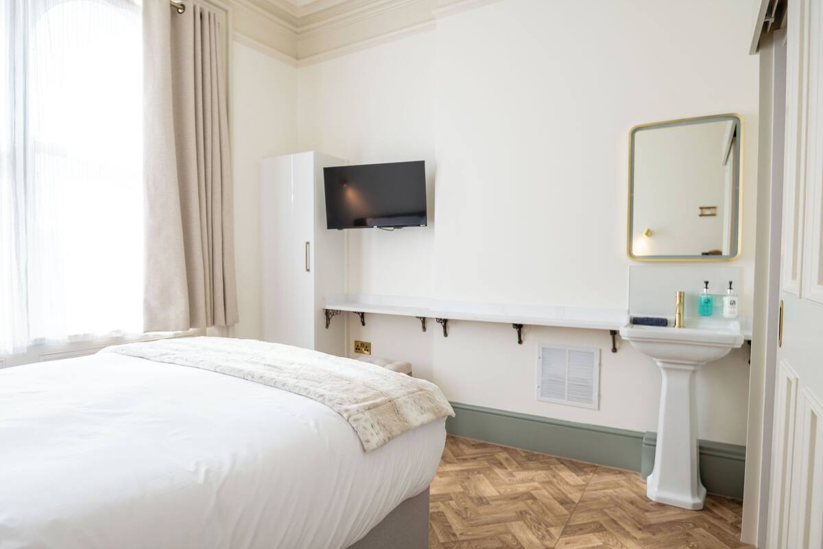 A hotel room with a bed, a mounted TV, a vanity with a mirror, and a sink.