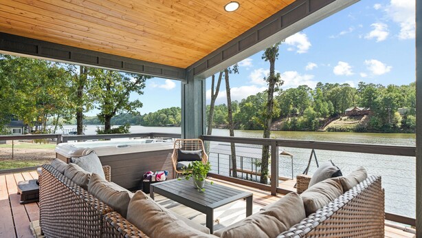 A covered deck with wicker furniture, a table with a plant, and a view of a lake and forest.
