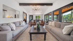 A spacious living room with two sofas, a coffee table, and a chandelier. There are large windows offering a view of the outdoors.