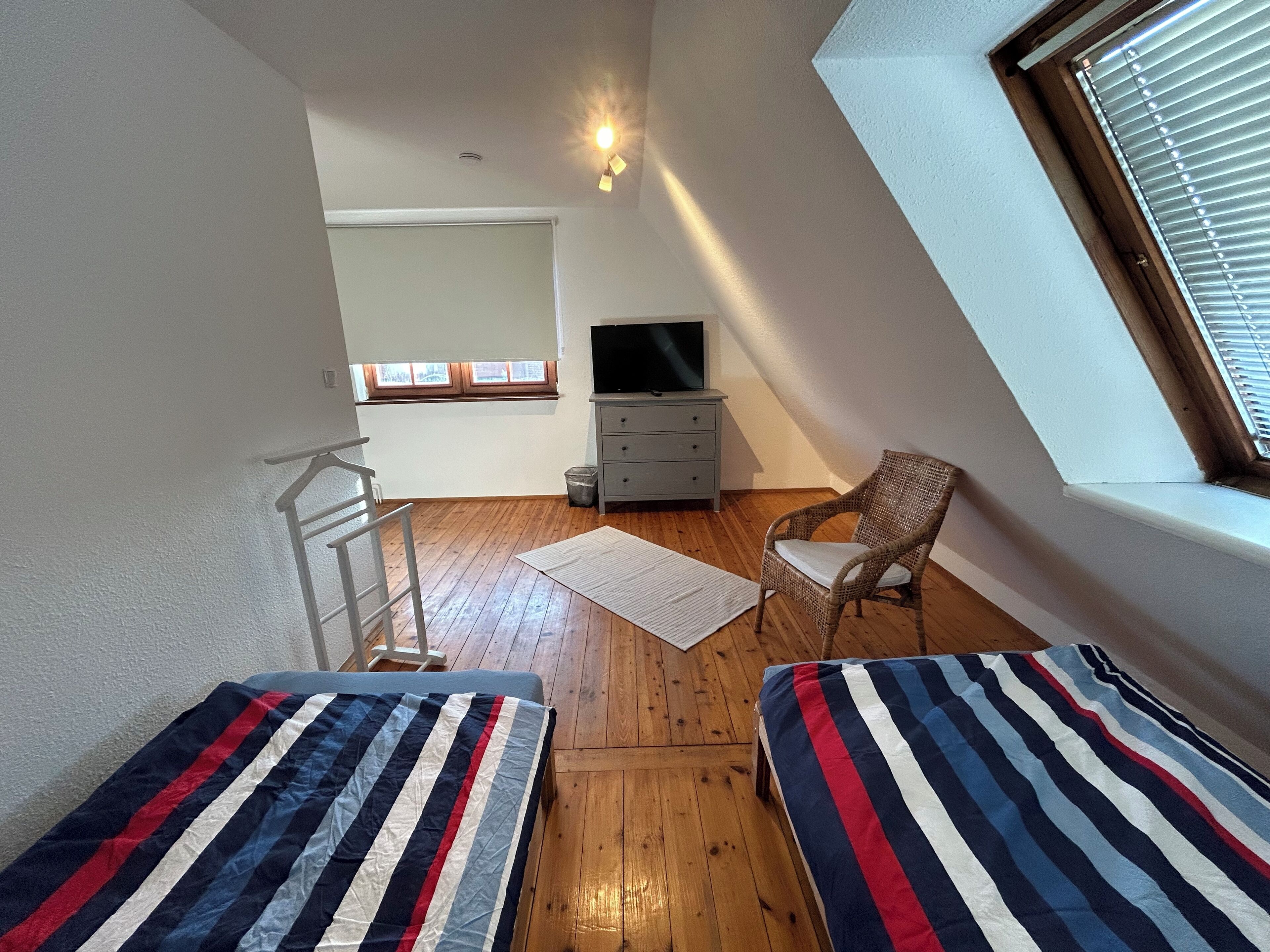 A small, cozy room with two single beds, a television, a chair, and a window with blinds.
