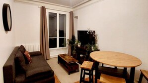 A living room with a dark-colored sofa, a wooden coffee table, a round wooden table with two chairs, a flat-screen TV, and a window with curtains. - Large, charming apartment near Montparnasse (Paris)