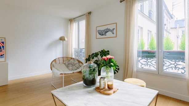 A modern living room with a large window, a dining table with a vase of flowers, a wicker chair, and a floor lamp. - Charming quiet apartment in the heart of Paris (Paris)