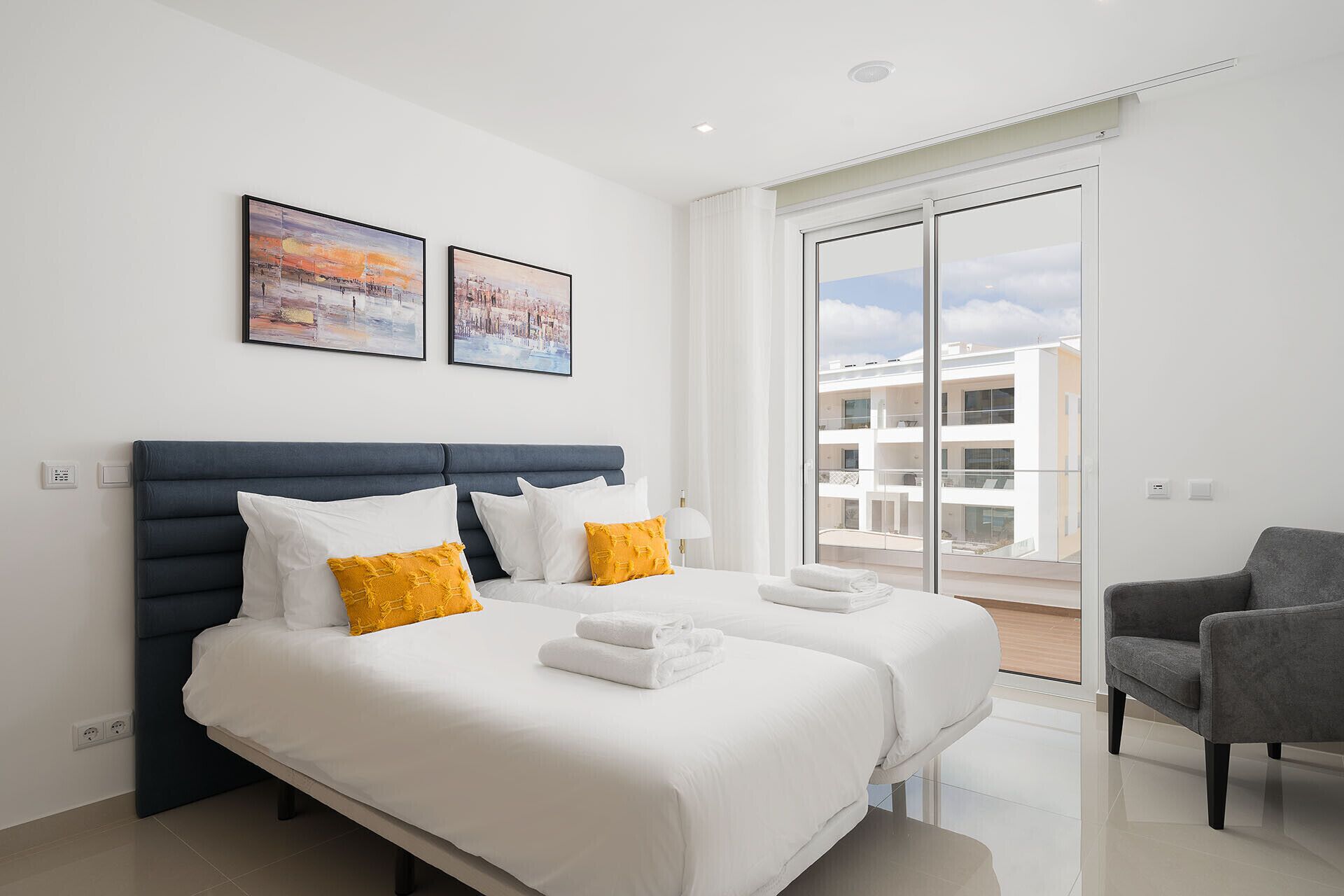 Une chambre d’hôtel moderne avec un grand lit, deux oreillers, une chaise et un balcon offrant une vue sur les immeubles.
