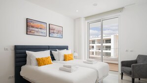 Habitación de hotel moderna con una cama grande, dos almohadas, una silla y un balcón con vistas a edificios.