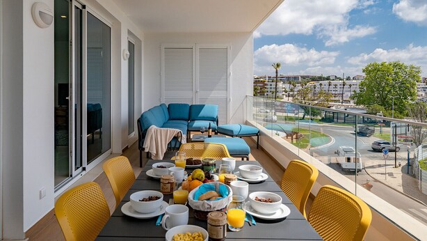Un balcón con una mesa de desayuno, un sofá azul y vistas a un paisaje urbano con edificios y una carretera.