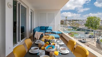 Un balcon avec une table à manger dressée pour le petit-déjeuner, un canapé bleu et une vue sur un paysage urbain avec des bâtiments et une route.