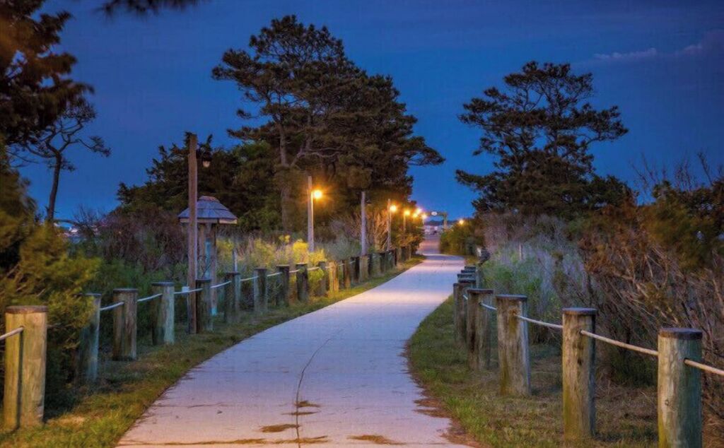 Un sendero pavimentado bordeado por postes de madera y un farol, que conduce hacia una edificación a lo lejos.