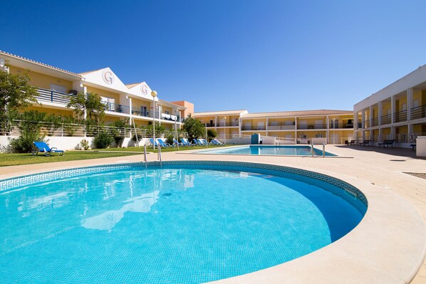 A swimming pool with a tiled edge, surrounded by a building with balconies and a clear blue sky.