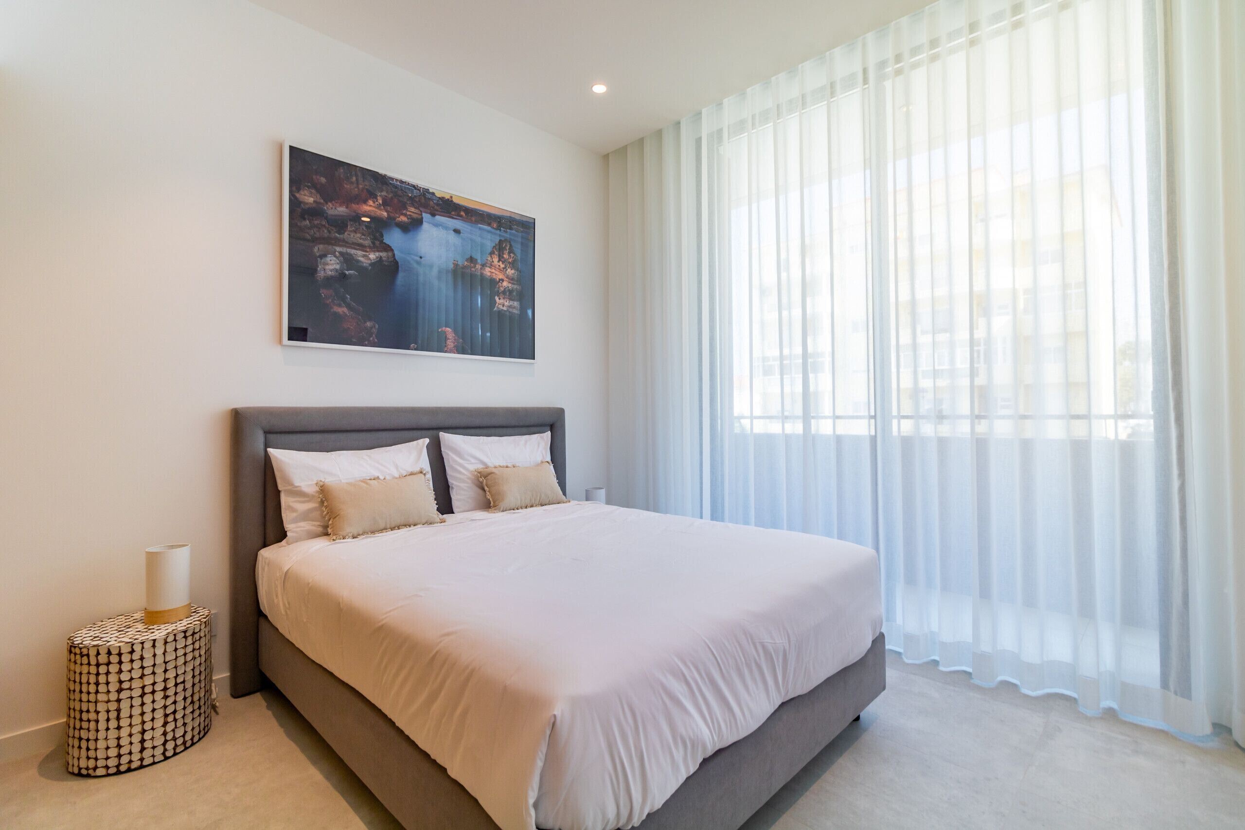 A modern bedroom with a large bed, a nightstand, a painting, and sheer curtains.