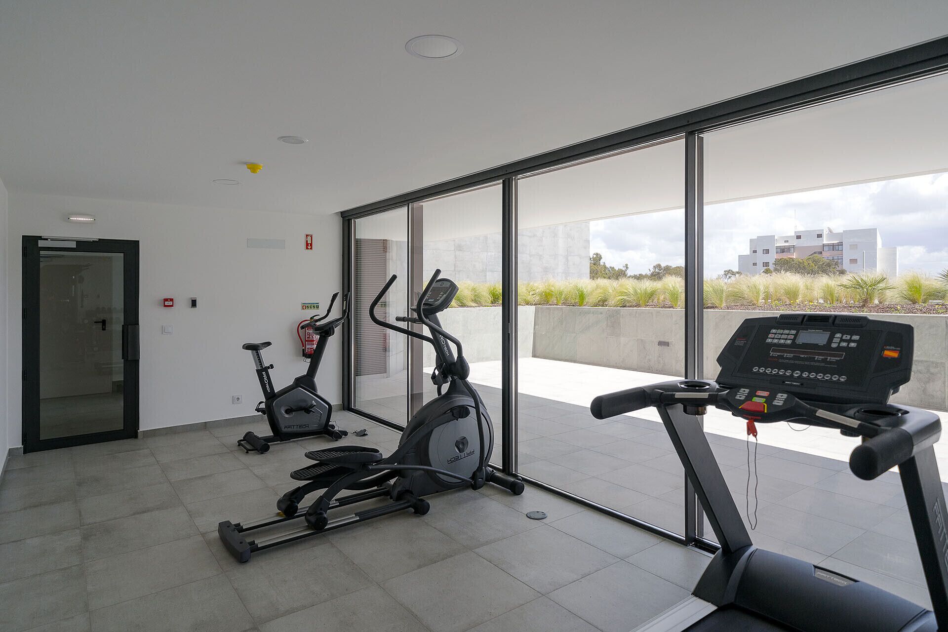 A modern gym with two stationary bikes and a treadmill, large windows offering a view of the city.