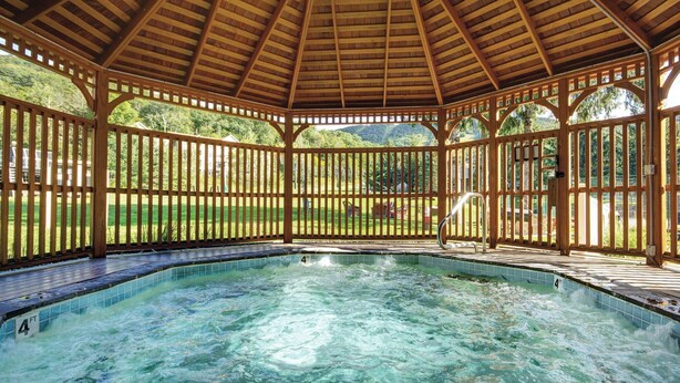 A hot tub area with a wooden gazebo structure, surrounded by a fence, and a view of a lush green landscape.