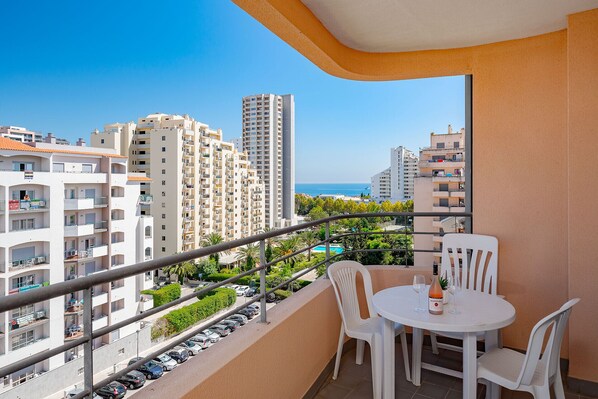 A balcony with a table and chairs, overlooking a parking area and a pool.