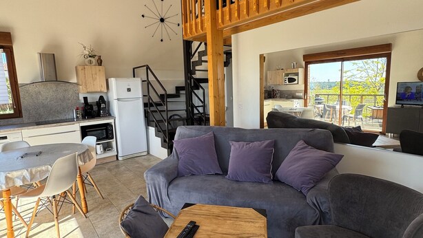 A spacious living area with a grey sofa, a wooden coffee table, and a dining table with chairs. A kitchen is visible in the background with a microwave and a refrigerator. - Chalet on the hill (Calvisson)