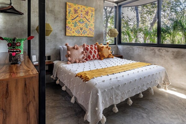 A bedroom with a large bed, a colorful abstract painting, and a view of trees through the window. - Architect-Designed 4BR Villa · Gated · Private Pool & Gym | Aldea Zama, Tulum (Tulum)