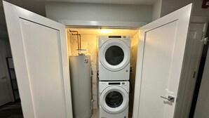 A compact laundry room with a front-loading washer and dryer, a water heater, and a door leading to another room. - The Comfy Suite: 1BR with Free Parking (Paterson)