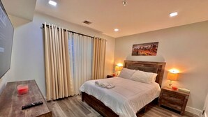 A hotel room with a wooden bed, a nightstand with a lamp, a TV on a wooden desk, and a cityscape painting on the wall. - The Comfy Suite: 1BR with Free Parking (Paterson)