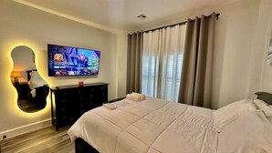 A modern hotel room with a large bed, a flat-screen TV mounted on the wall, a dark wooden dresser, and a unique wall mirror. - The Stylish Spot: 1BR with Free Parking (Paterson)