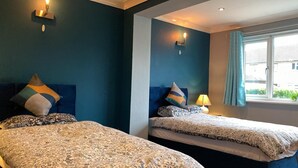 Ein Hotelzimmer mit zwei Betten, jedes mit gemusterten Bettbezügen und dekorativen Kissen. An den Wänden sind Lampen befestigt und ein Fenster mit Vorhängen vorhanden.