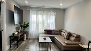 A living room with a brown leather sofa, a small coffee table, a TV mounted on the wall, and a window with sheer curtains. - The Lovely Suite: 1BR close to NYC (Paterson)